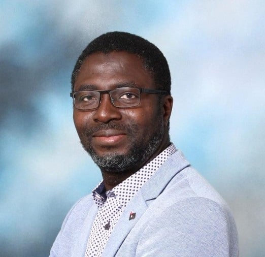 Man with glasses and beard wearing a light gray blazer over a polka dot shirt, against a blue gradient background.