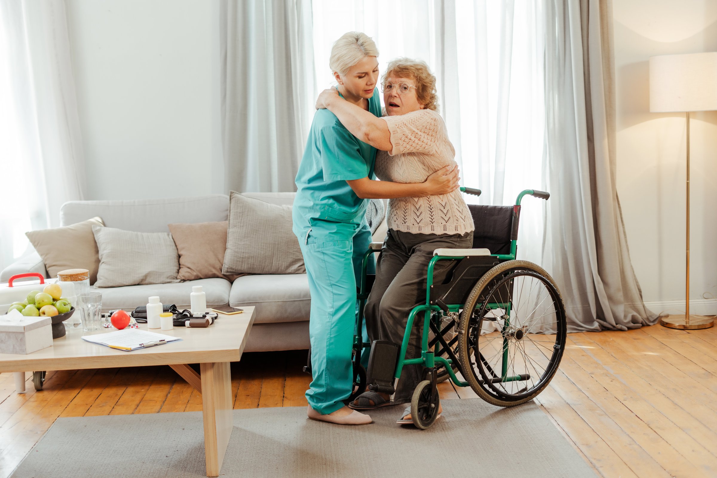 Young nurse supporting a senior woman while she is struggling to get up from the wheelchair in a home health care concept