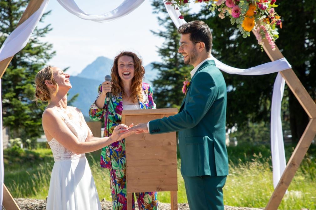 A bride and groom exchange vows outdoors, officiated by a smiling woman. Decor includes floral arrangements and white drapes.