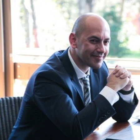 A bald man in a suit smiles while seated at a table, hands clasped, with a window and greenery in the background.