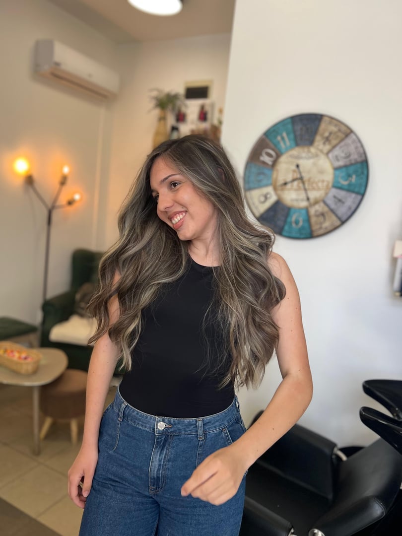 Woman with long wavy hair, wearing a black top and jeans, smiling in a salon with a vintage clock on the wall behind her.