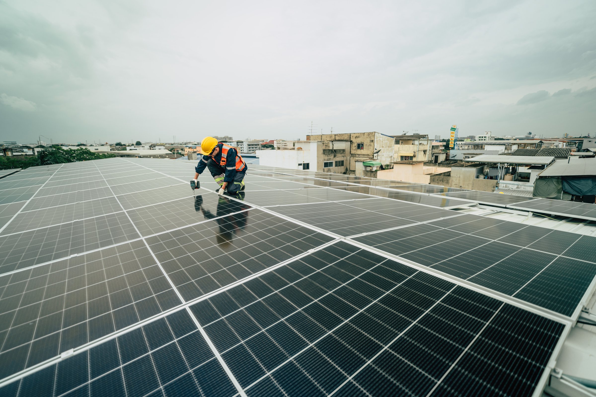 A technician carefully installs solar panels on a rooftop, surrounded by a bustling urban landscape. The cloudy sky hints at possible rain while the city lies beneath.