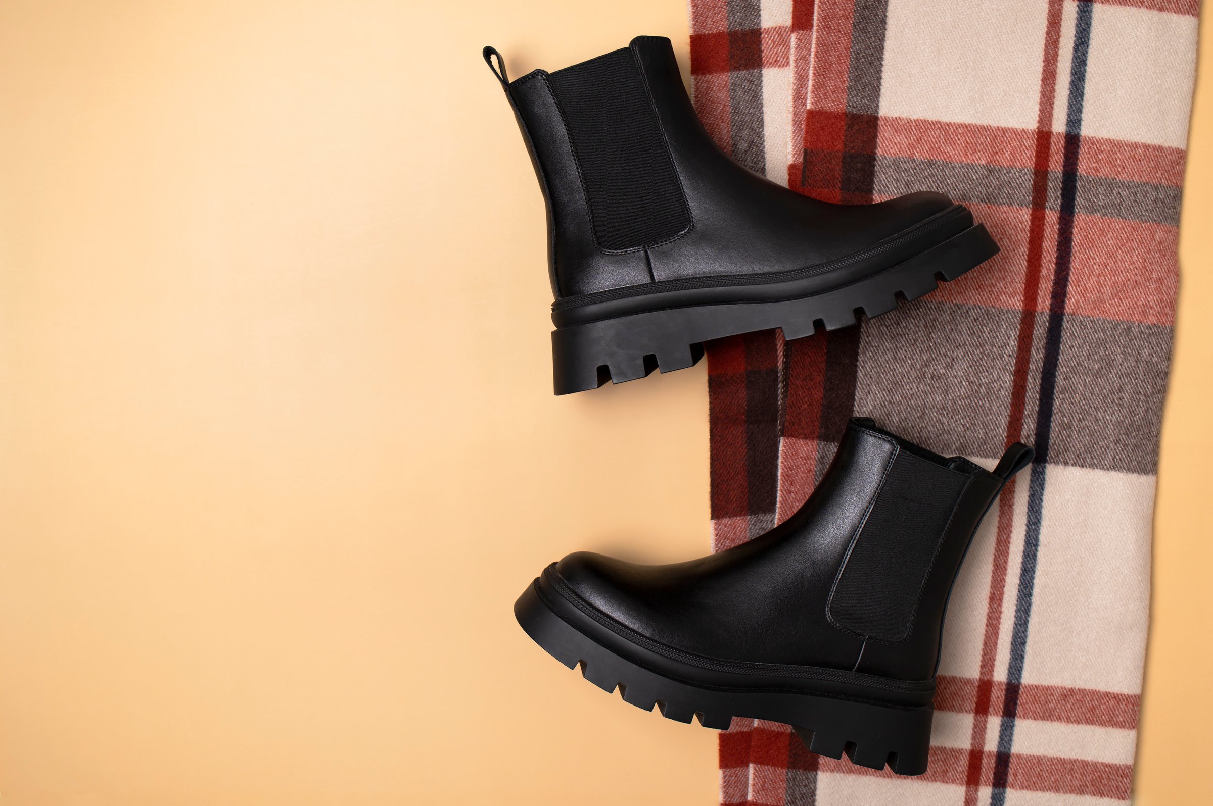 Black leather classic women's boots and checkered autumn wool scarf on beige background top view flat lay. Minimalistic layout, autumn footwear advertisement. Fashionable stylish boots
