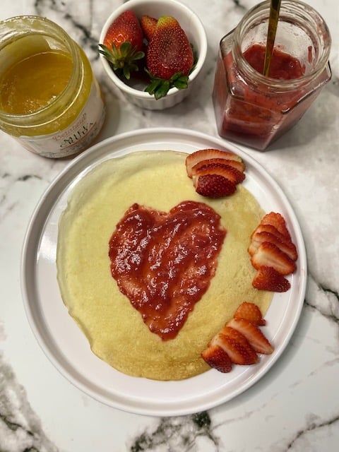Crepe with heart-shaped strawberry jam, sliced strawberries, honey jar, and bowl of strawberries on a marble surface.
