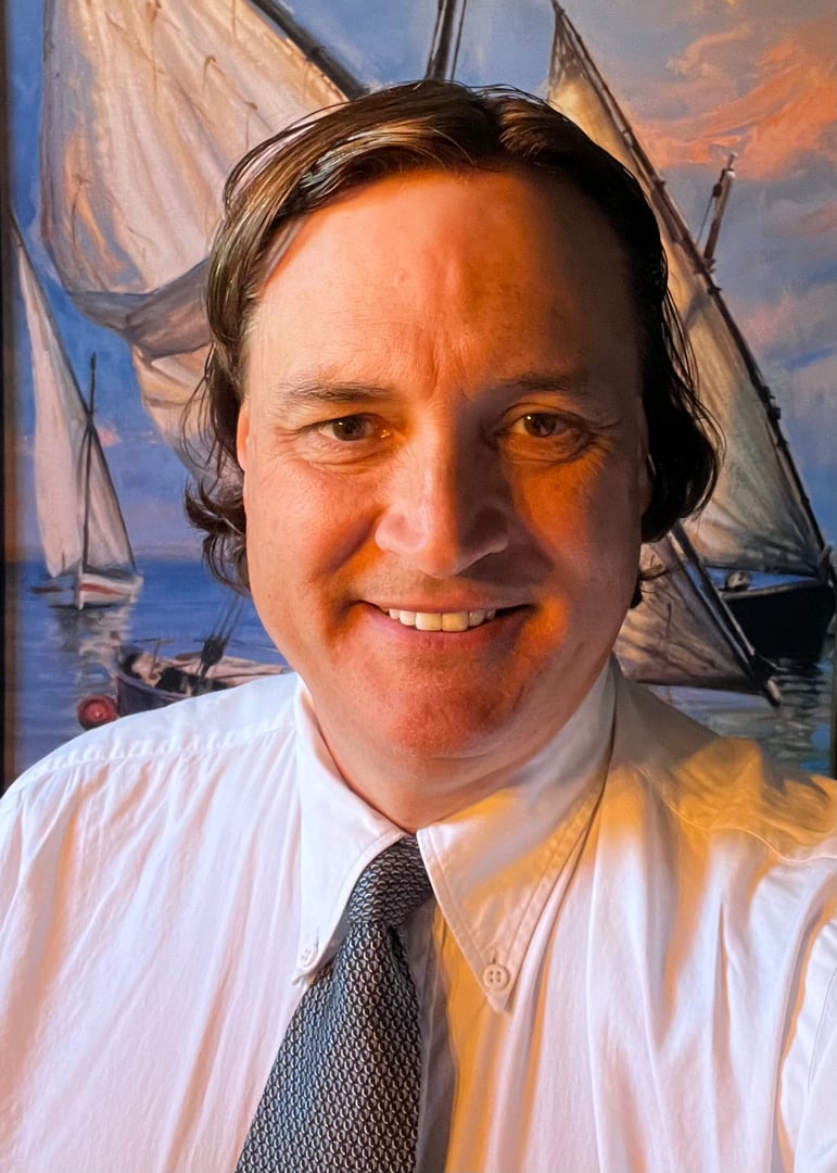Man in a white shirt and tie smiles in front of a painting of sailboats on a sunny day.