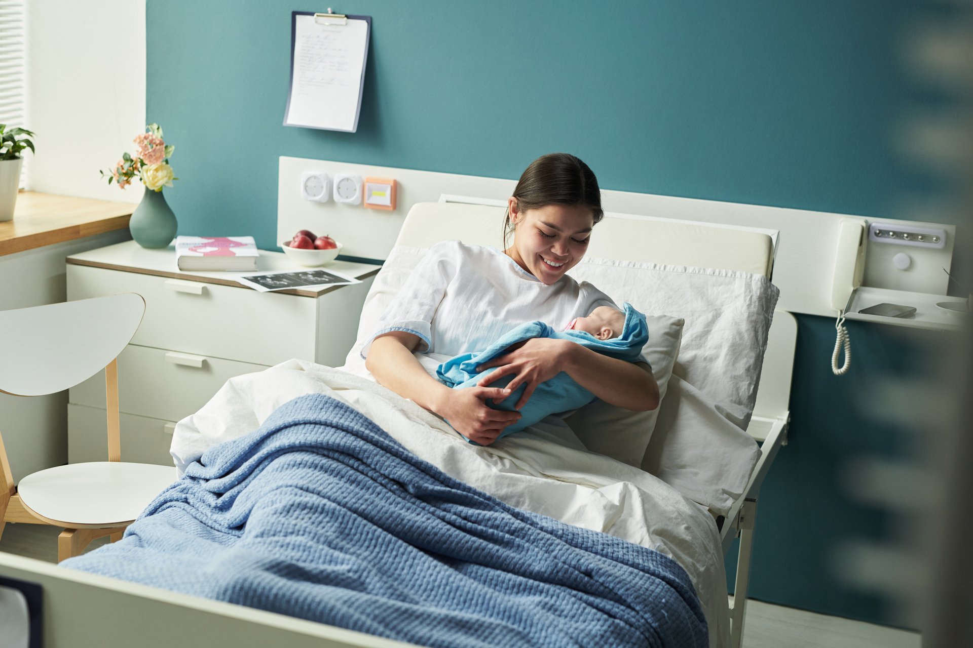 Young Caucasian woman lying in hospital bed holding newborn baby, smiling and looking at infant, modern medical room background, mother bonding with child after birth