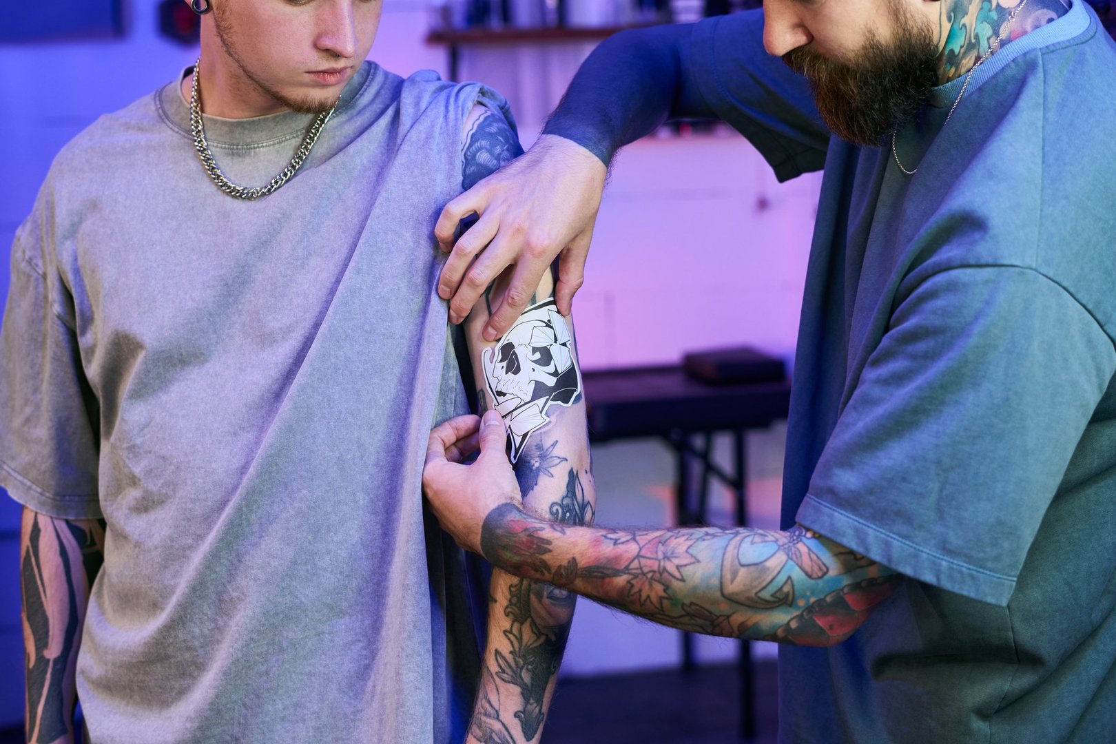 Tattoo artist placing detailed skull stencil on client arm while preparing for session inside professional studio with cool-toned ambient lighting and focused setting