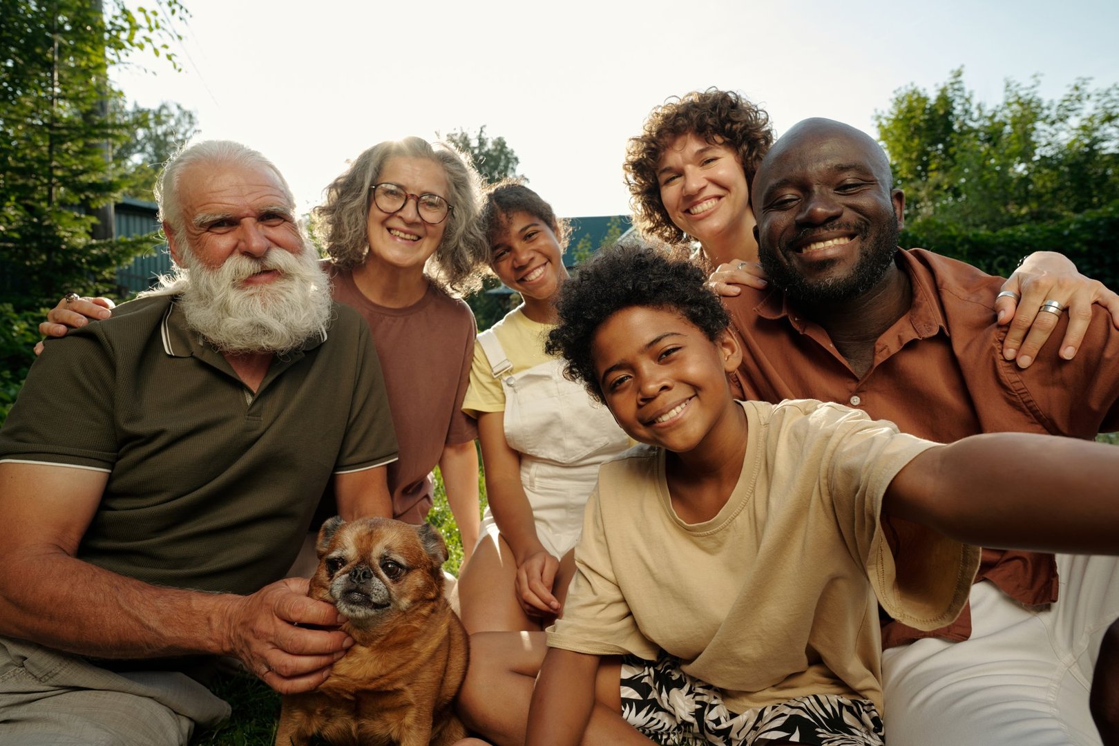 Happy intercultural family of three generations sitting on green lawn in natural environment and making selfie while enjoying being together