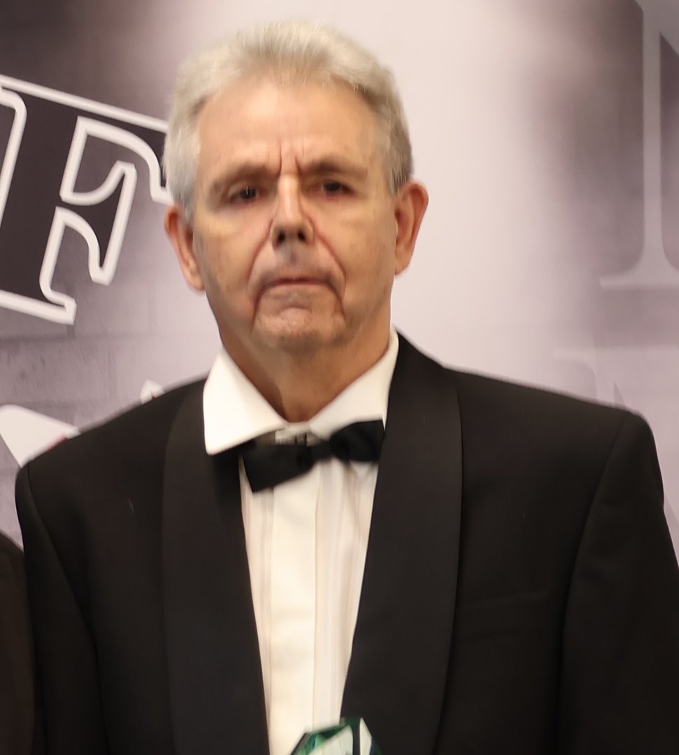 Elderly man in a tuxedo with a bow tie, holding a trophy. Background features blurred letters on a wall.