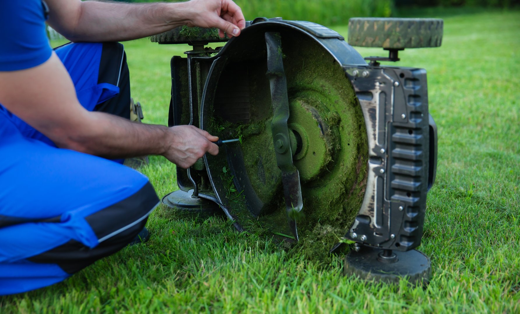 Man in blue overalls is carefully adjusting a lawn mower blade and cleaning while kneeling on a well-kept lawn. The importance of proper equipment maintenance for effective lawn care.