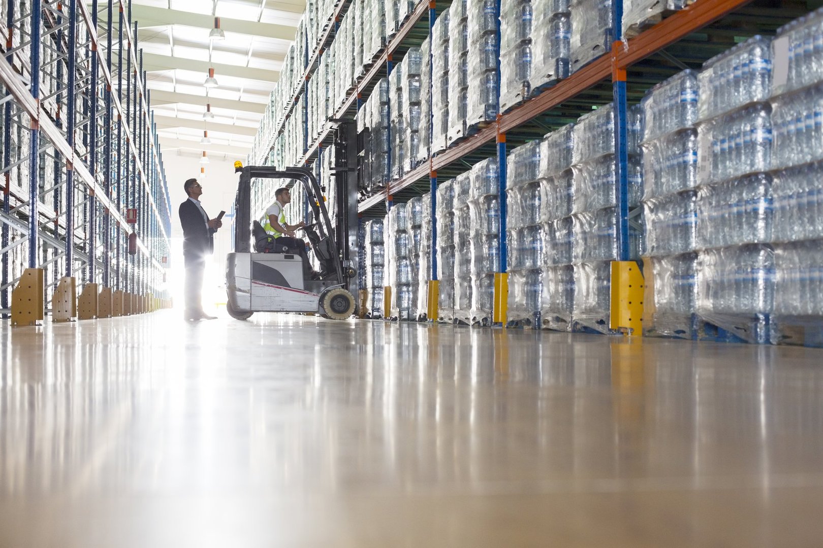 Worker operating a forklift in a warehouse filled with stacked water bottles while a man observes, sunlight shining in.