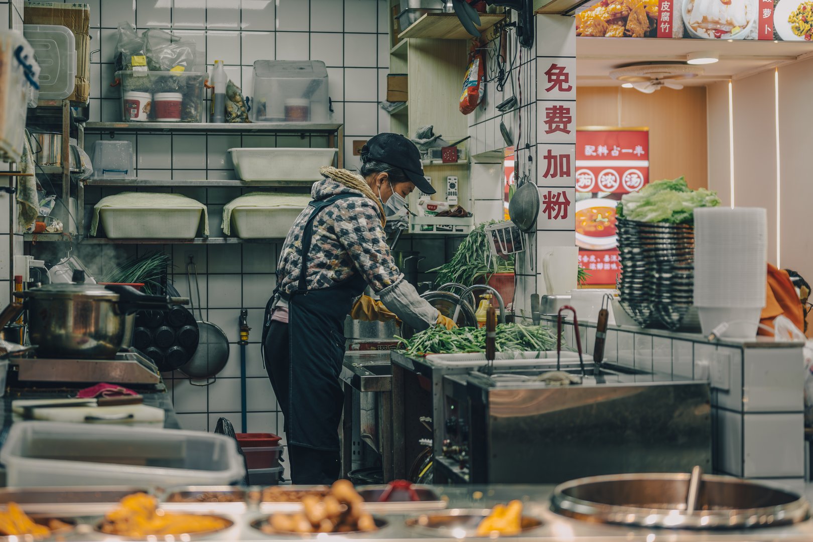 Guangzhou, Canton, China, 2 March 2025: woman washing vegetables in a small kitchen of local restaurant with tiled walls and steam rising from pots. Everyday culinary life in southern China.