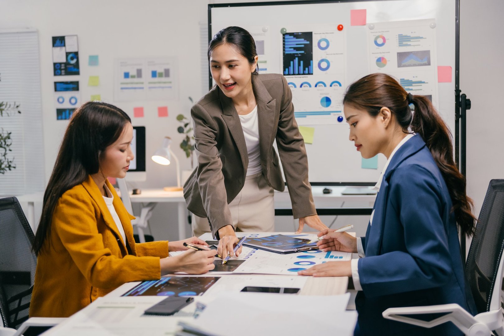 Three young asian businesswomen are meeting in a modern office, analyzing charts and graphs displayed on a whiteboard and spread out on the table, collaborating on a project