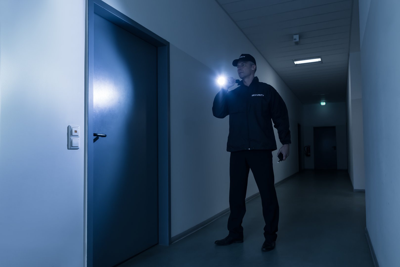Full length of mature security guard with flashlight standing in front of door in building