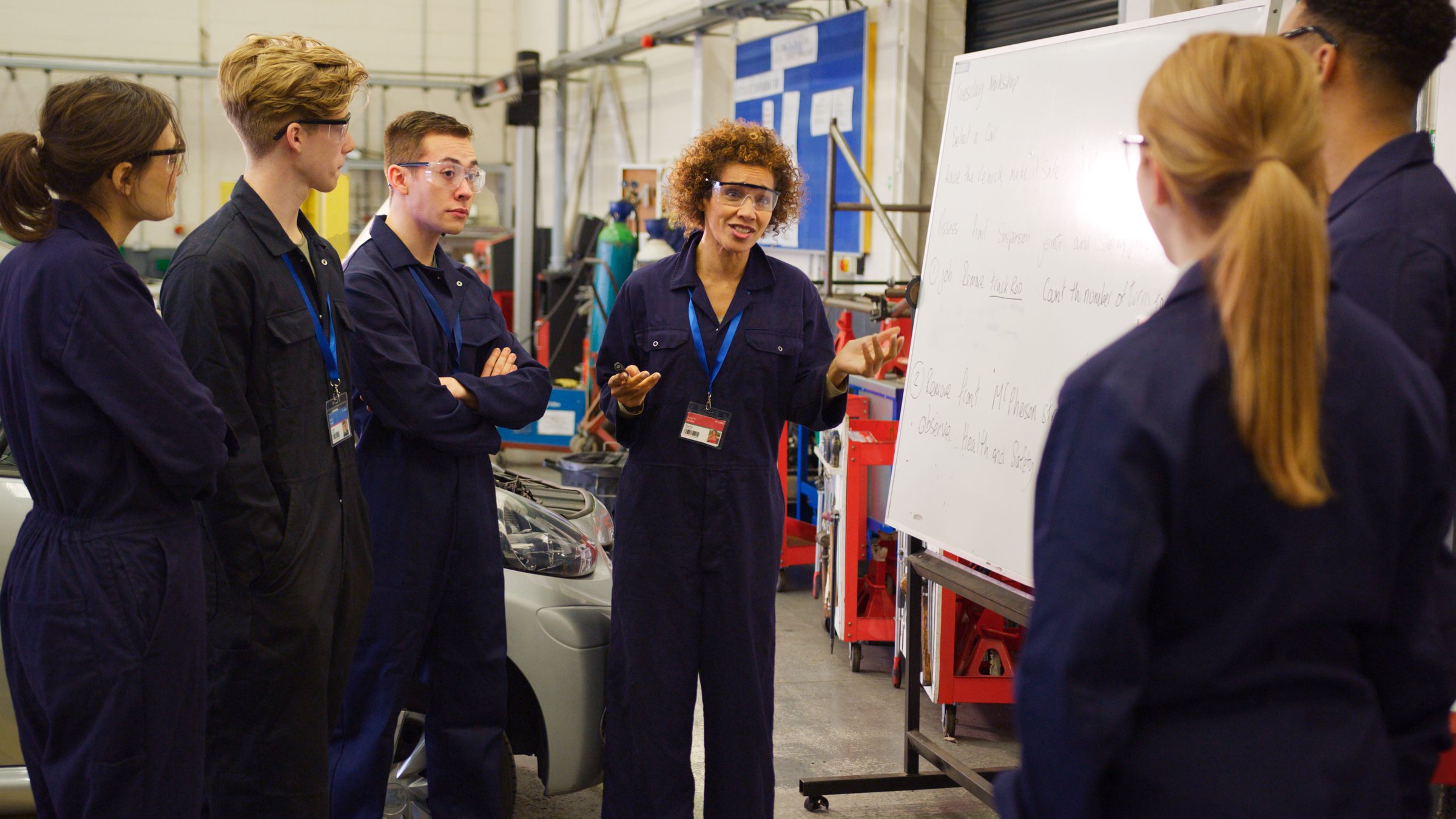 Female Tutor By Whiteboard With Students Teaching Auto Mechanic Apprenticeship At College
