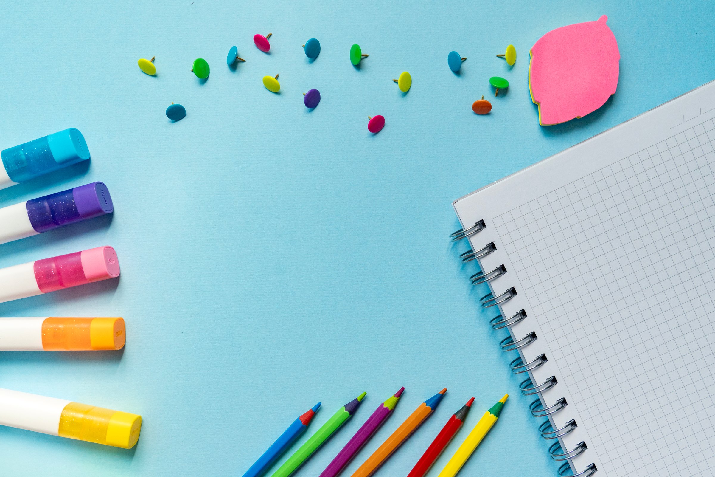 Top view of colorful stationery including notebook, markers, pencils, and sticky notes arranged on blue background.