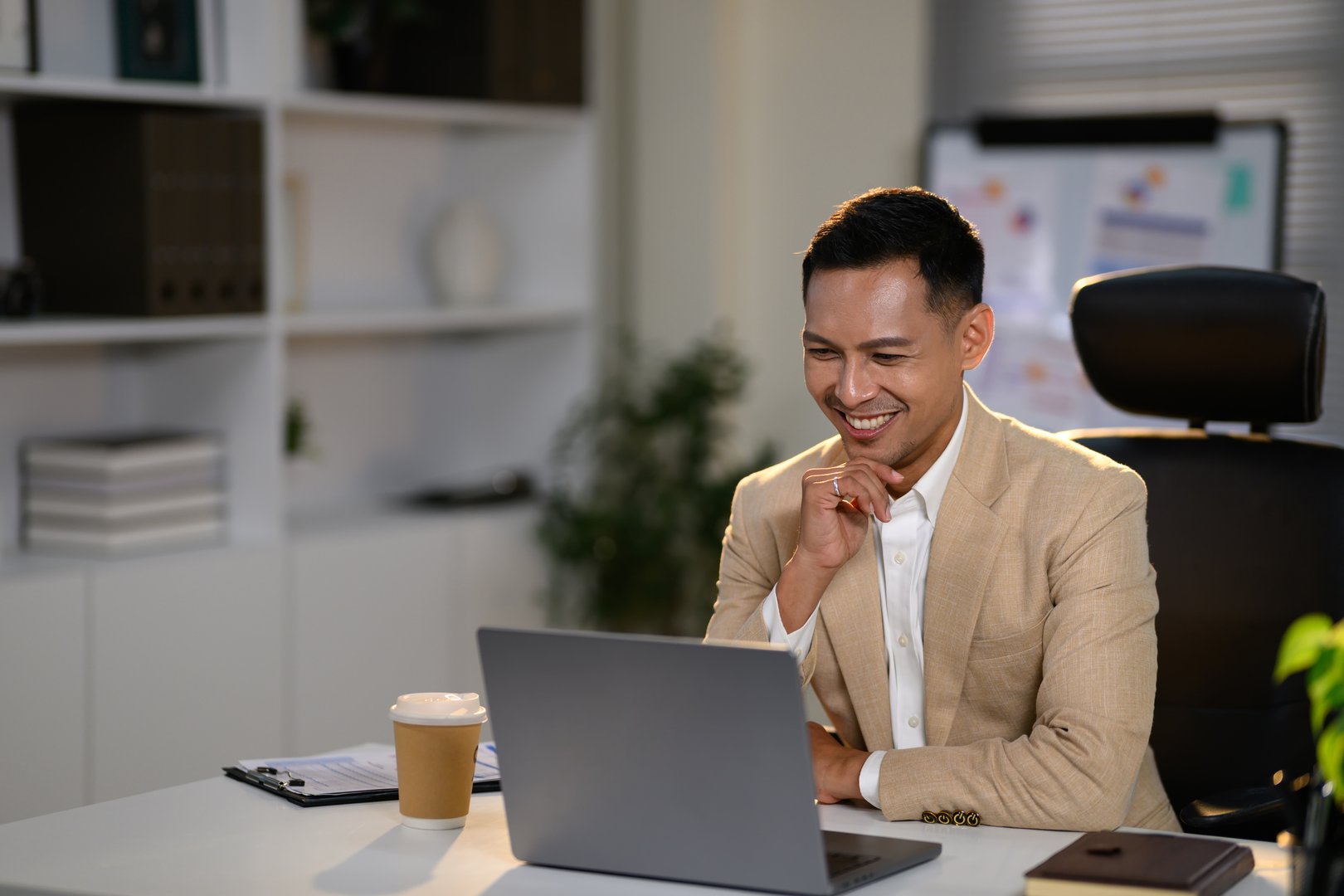 Confident businessman in a beige suit smiling, attending an online meeting at his desk.