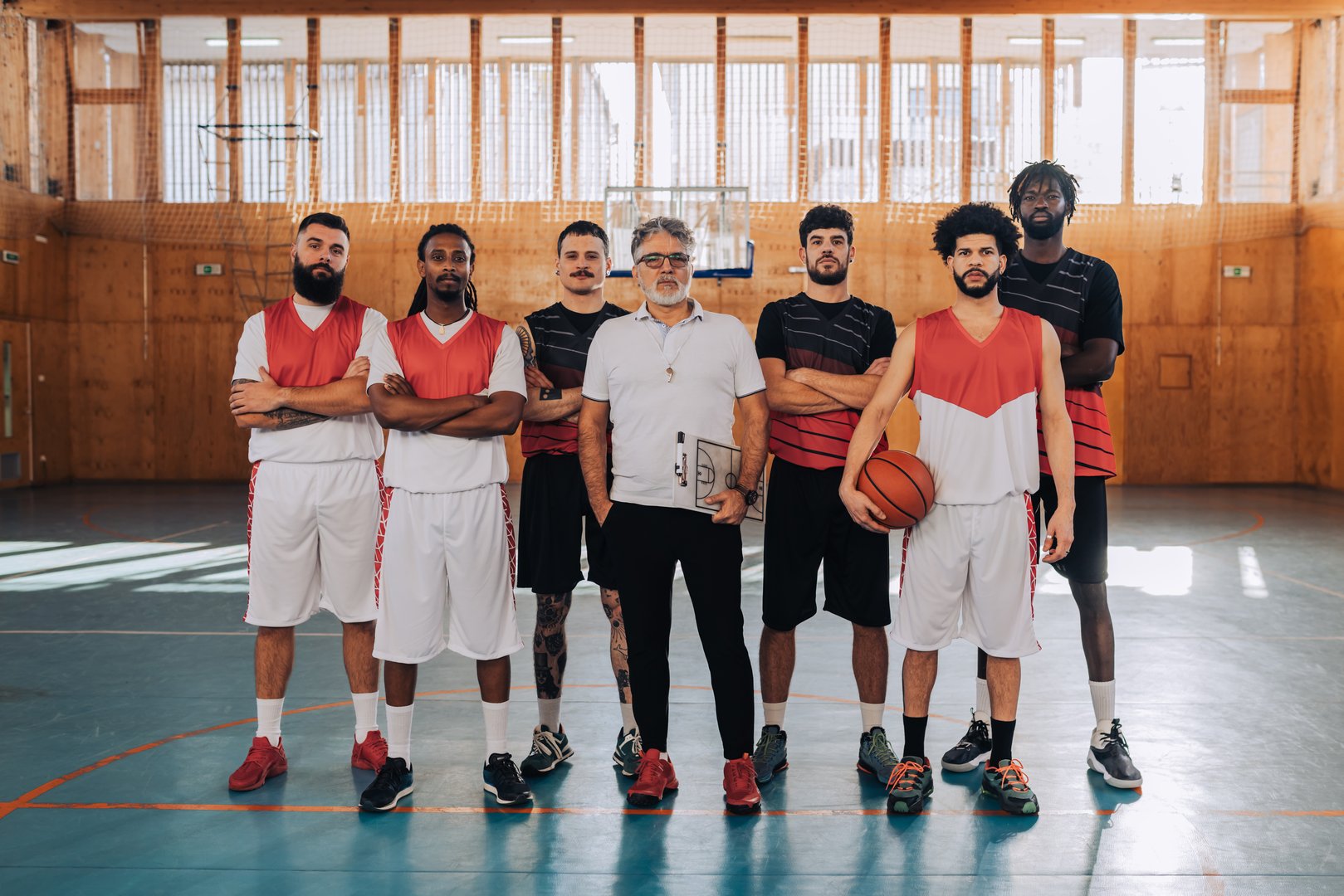 Portrait of a diverse basketball team and their elderly coach standing on a court in an indoor sports hall and posing while looking at the camera before training or a game. Diversity in sport.