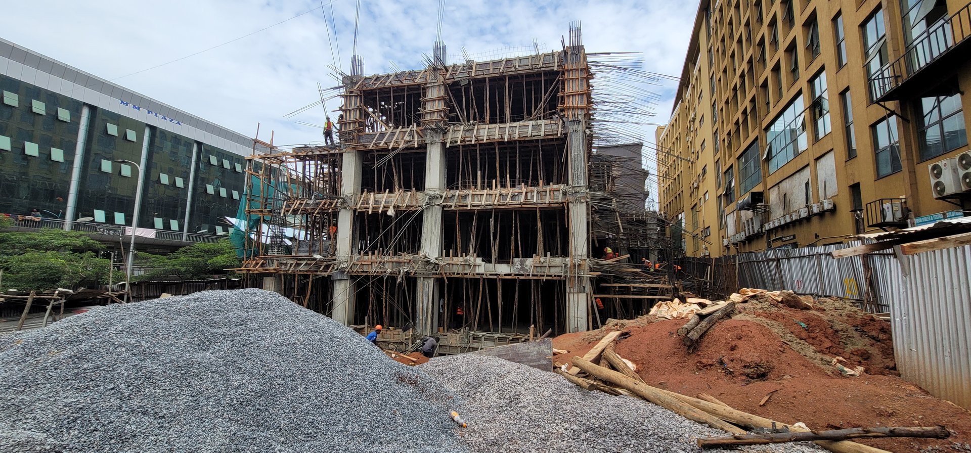 Construction site with a multi-story building frame, piles of gravel, and a nearby office building on a cloudy day.