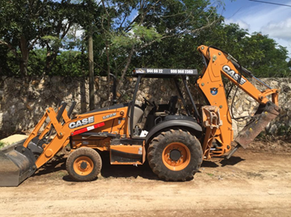 An orange CASE backhoe loader parked on a dirt road, surrounded by greenery and a stone wall in the background.