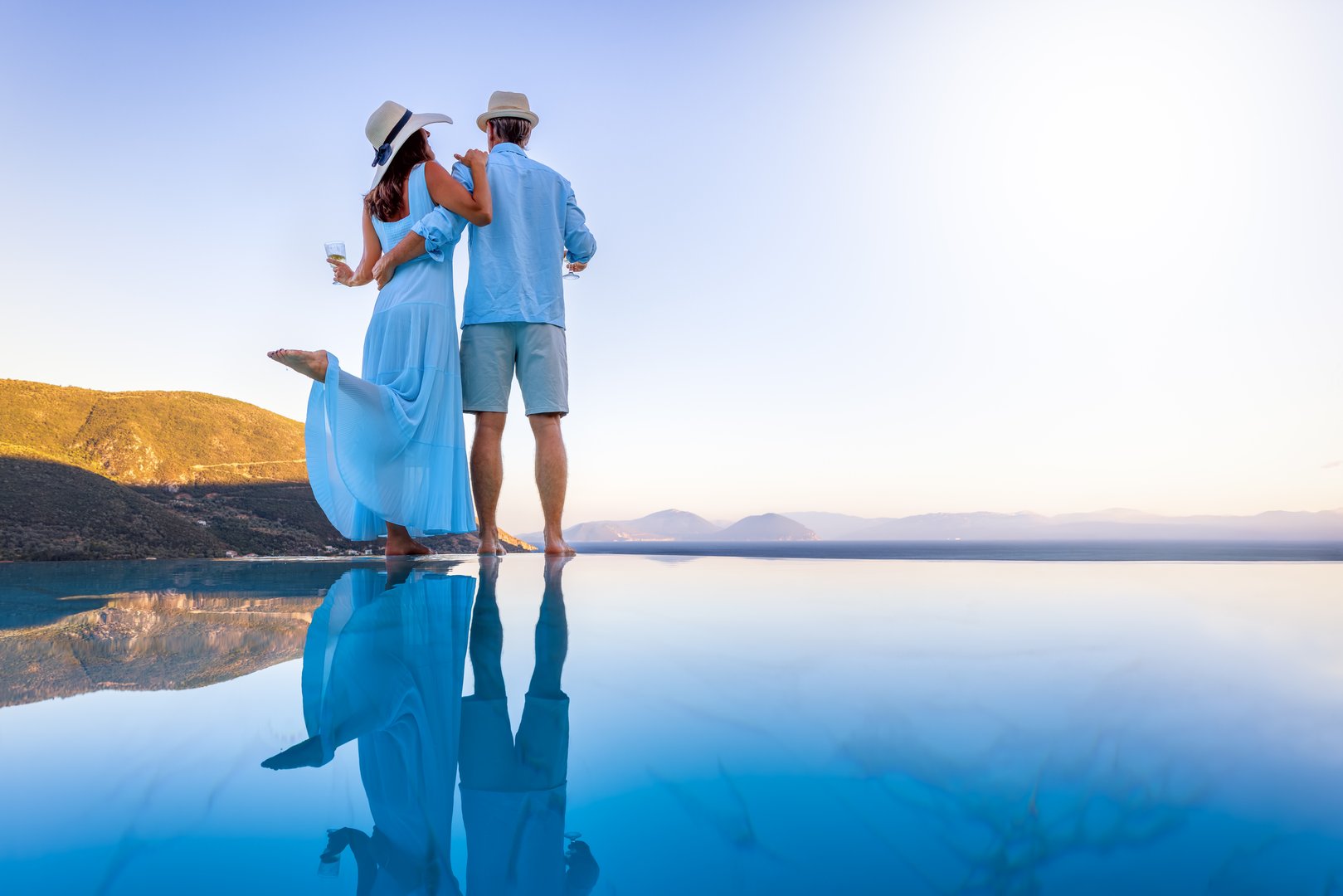 A elegant summer vacation or honeymoon couple enjoys the mediterranean sunset with a glass of wine by the pool