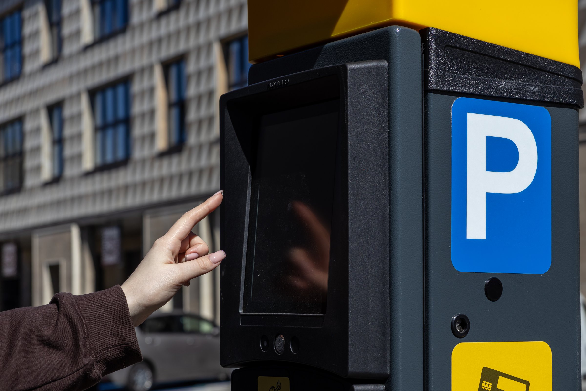 Close-up of a woman's hand interacting with a touchscreen parking meter on a city street. The modern terminal features a blue parking sign and payment options
