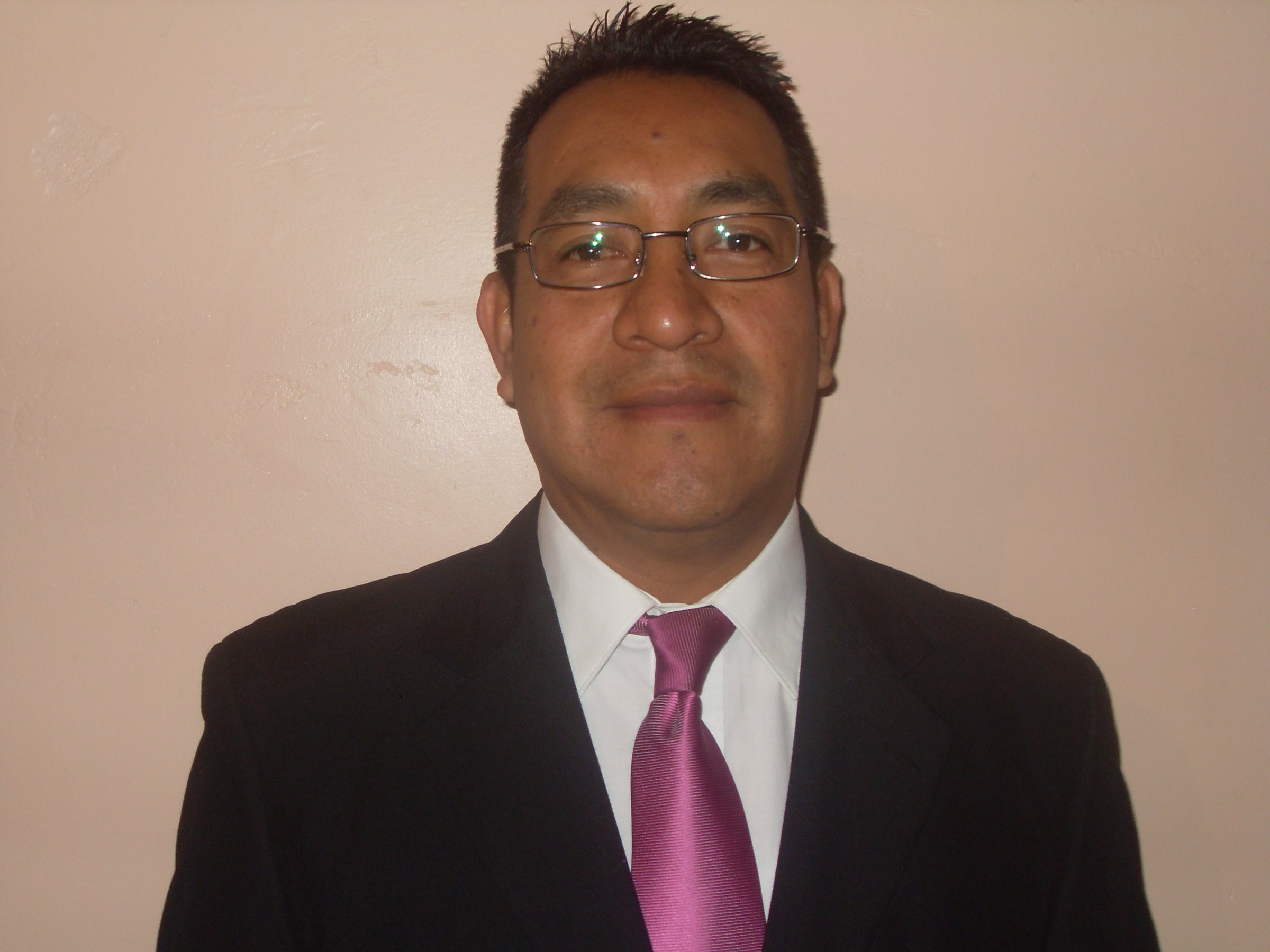 Man in a dark suit and purple tie against a light colored wall, wearing glasses, with short dark hair.