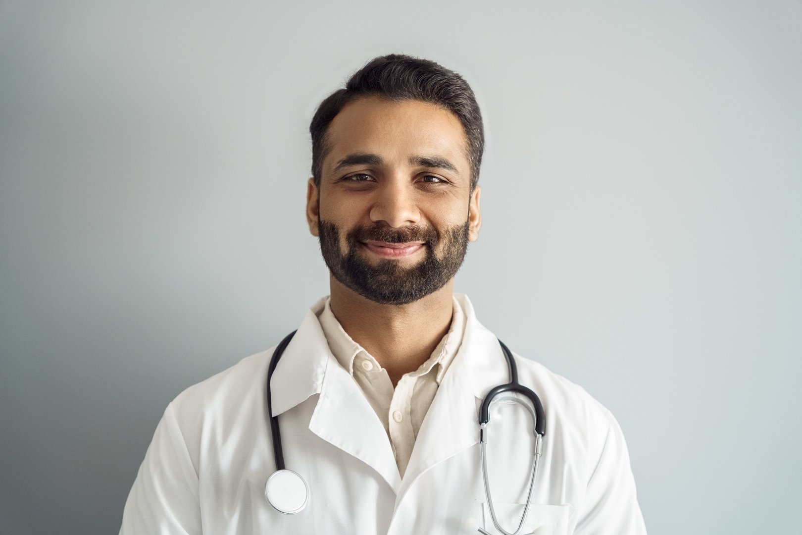 Headshot portrait of smiling doctor wearing white coat, stethoscope looking at camera isolated on white studio wall background. Therapist or general practitioner, medical staff, clinic presentation