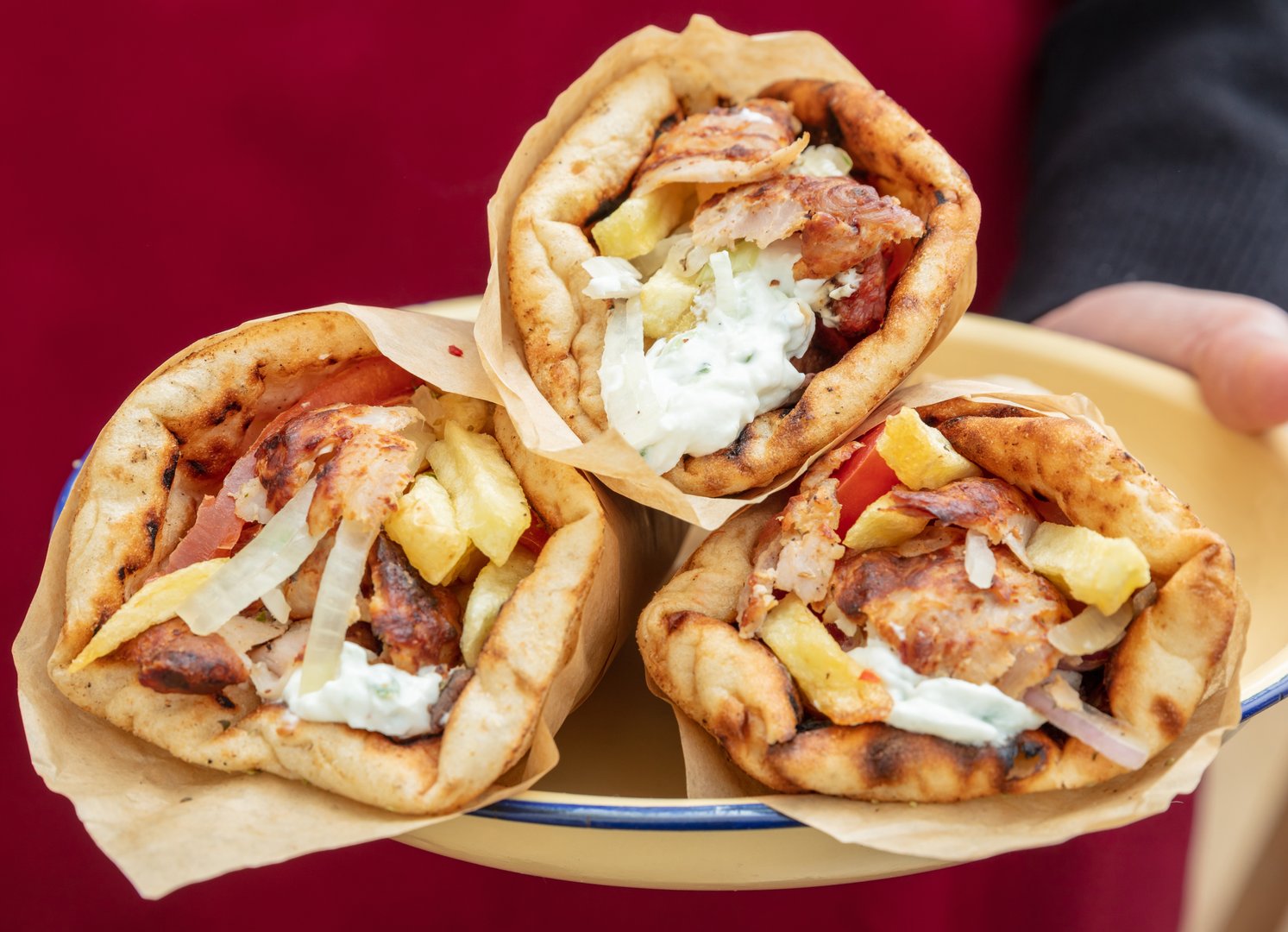Three freshly made Greek gyros wraps filled with grilled meat, fries, onions, and tzatziki sauce are served on a rustic plate against a warm red background in natural daylight.