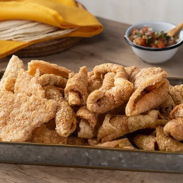 A tray of crispy pork rinds on a wooden table, with a bowl of salsa and a stack of tortillas in the background.