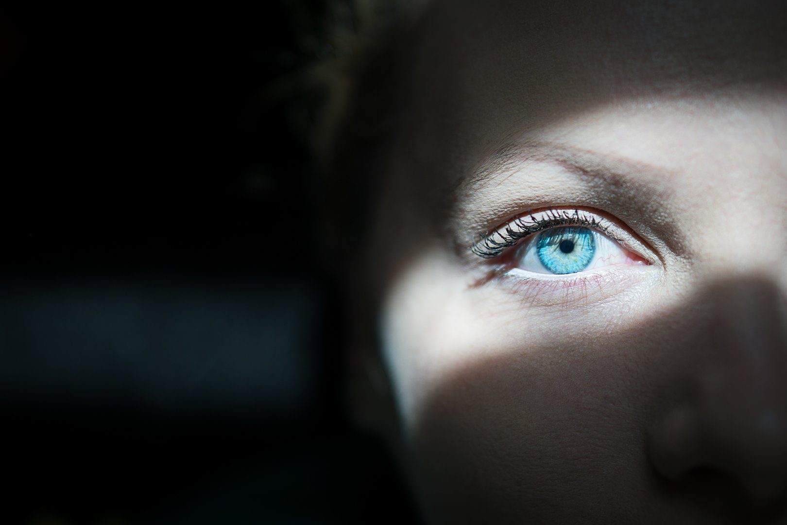 Close-up of a blue eye, partially illuminated by light, surrounded by shadow.