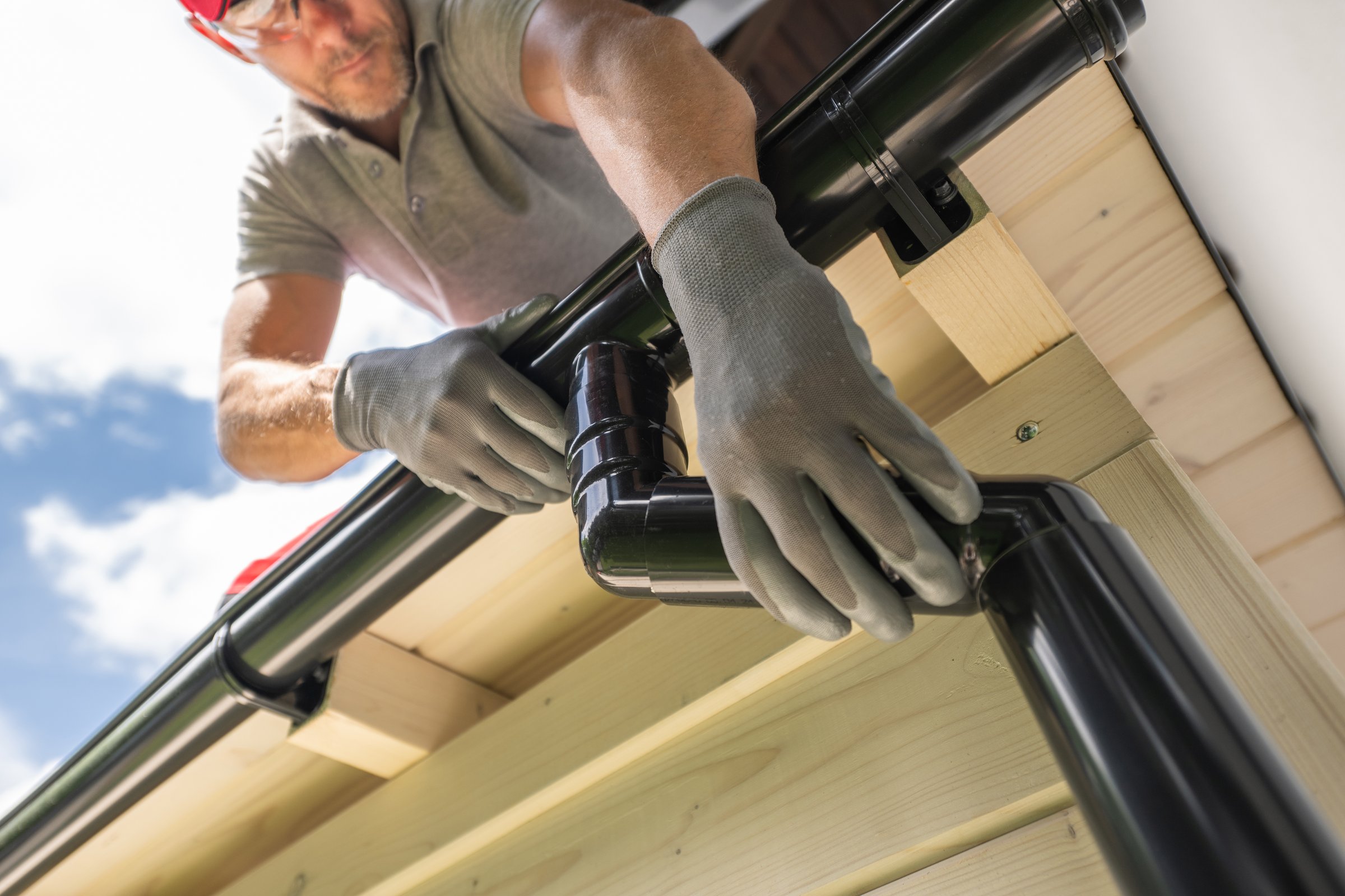 A worker wearing gloves and safety glasses installs a black gutter system on a residential roof. The worker is seen from below, looking up at the gutter system, which is attached to the wooden fascia board. The sky is visible in the background.