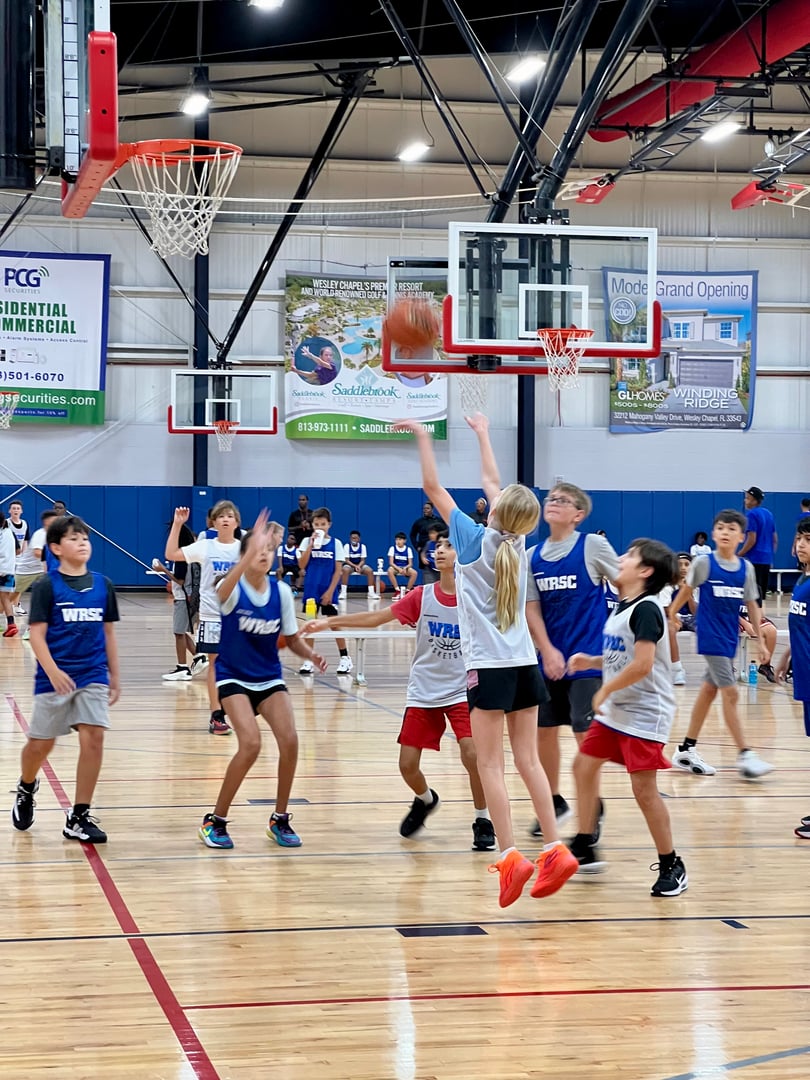 Children playing basketball indoors; one player shoots while others watch. The gym has banners and a hardwood floor.