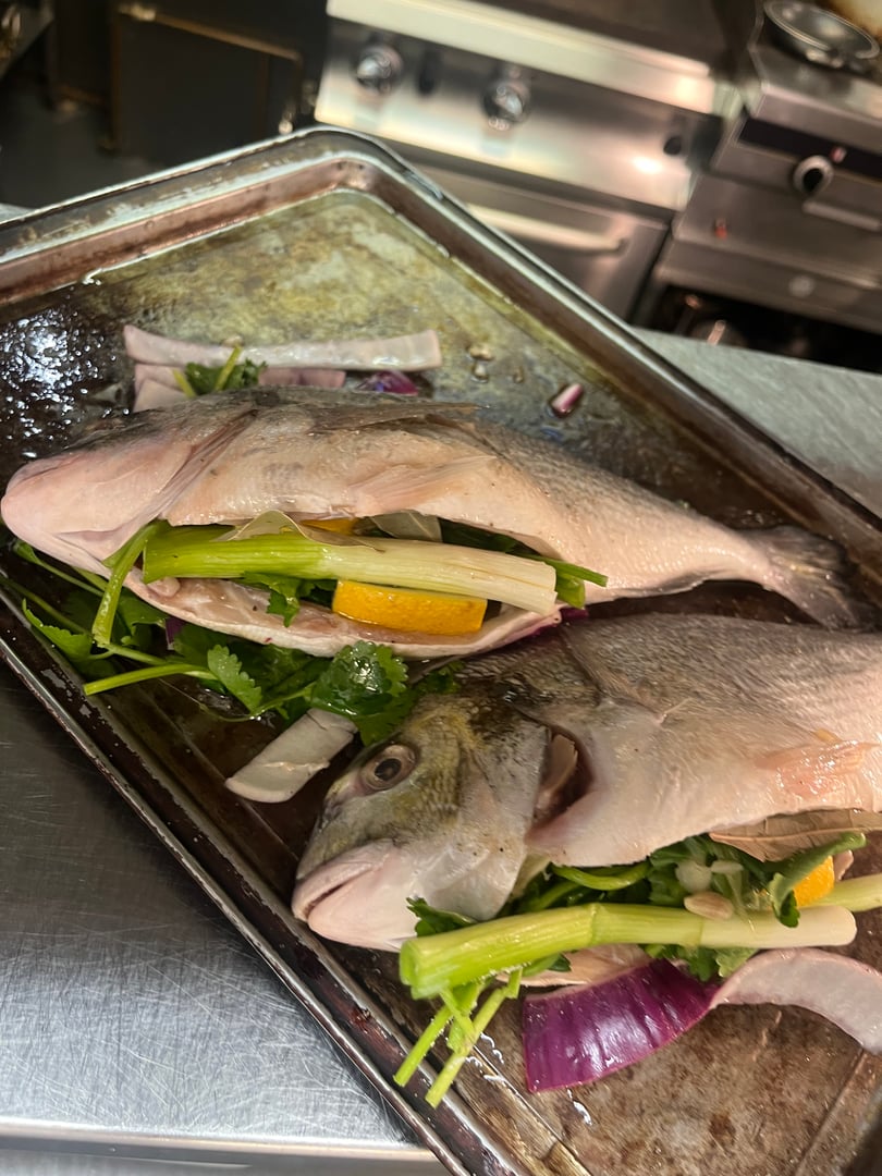 Two raw fish stuffed with celery, lemon, and herbs on a metal tray, ready for cooking in a kitchen setting.