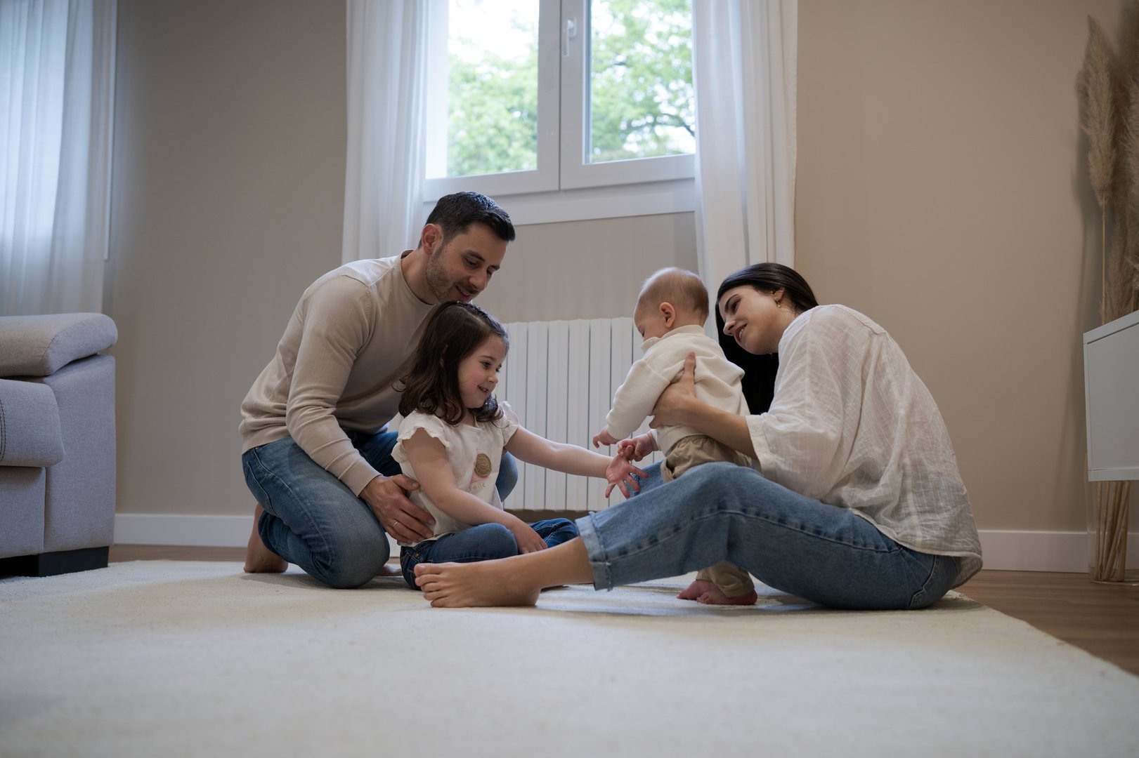 Young parents sitting on a cozy carpet with two children, a baby and a preschooler girl, enjoying playful moments together in a warm, inviting living room filled with natural light