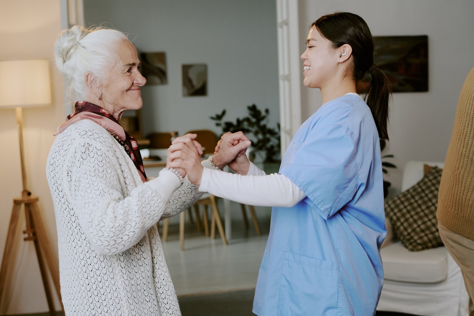 Senior Caucasian woman smiling while holding hands with young adult Hispanic female nurse, both standing and interacting in living room setting, showing supportive relationship