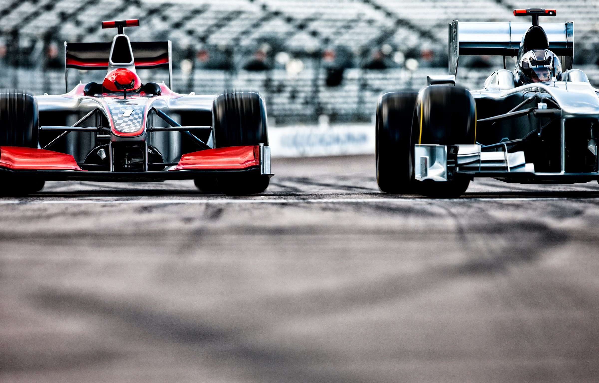 Two Formula 1 cars racing side by side on a track, with blurred stands in the background.