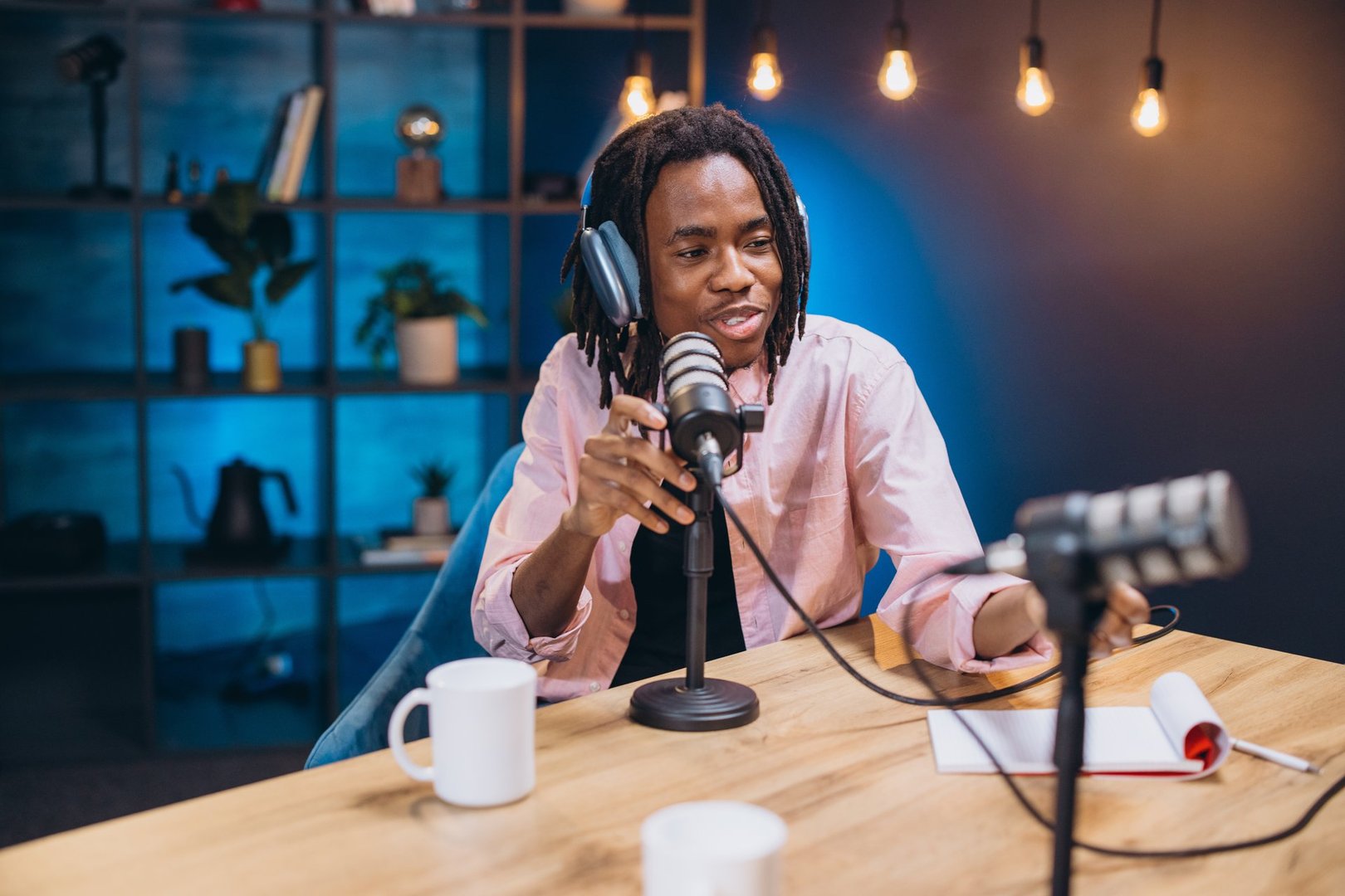 African American male professional content creator recording podcast, wearing headphones, speaking into microphone within contemporary broadcasting workspace