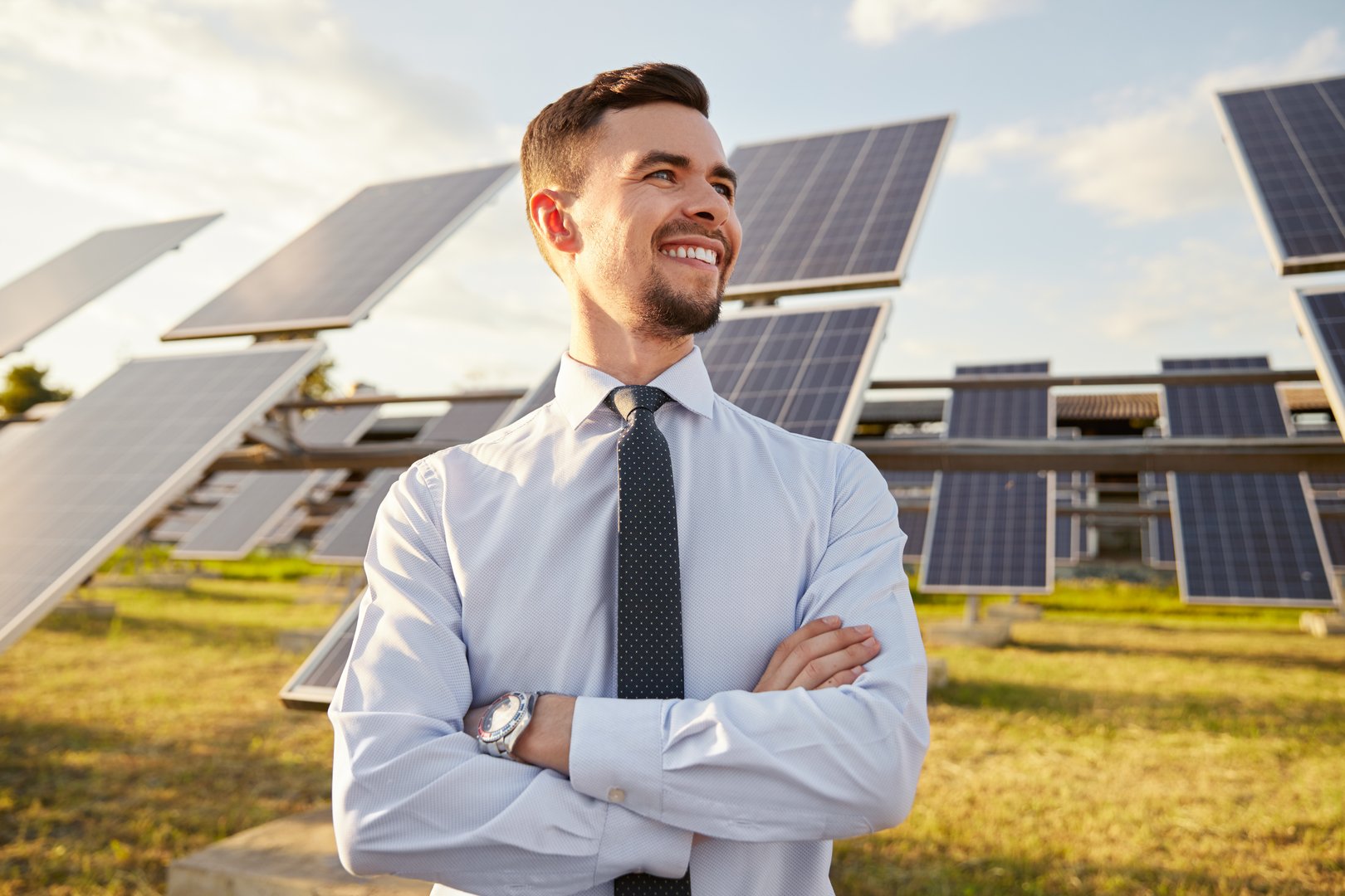 Positive male entrepreneur crossing arms and looking away with smile while standing near contemporary photovoltaic panels on solar farm