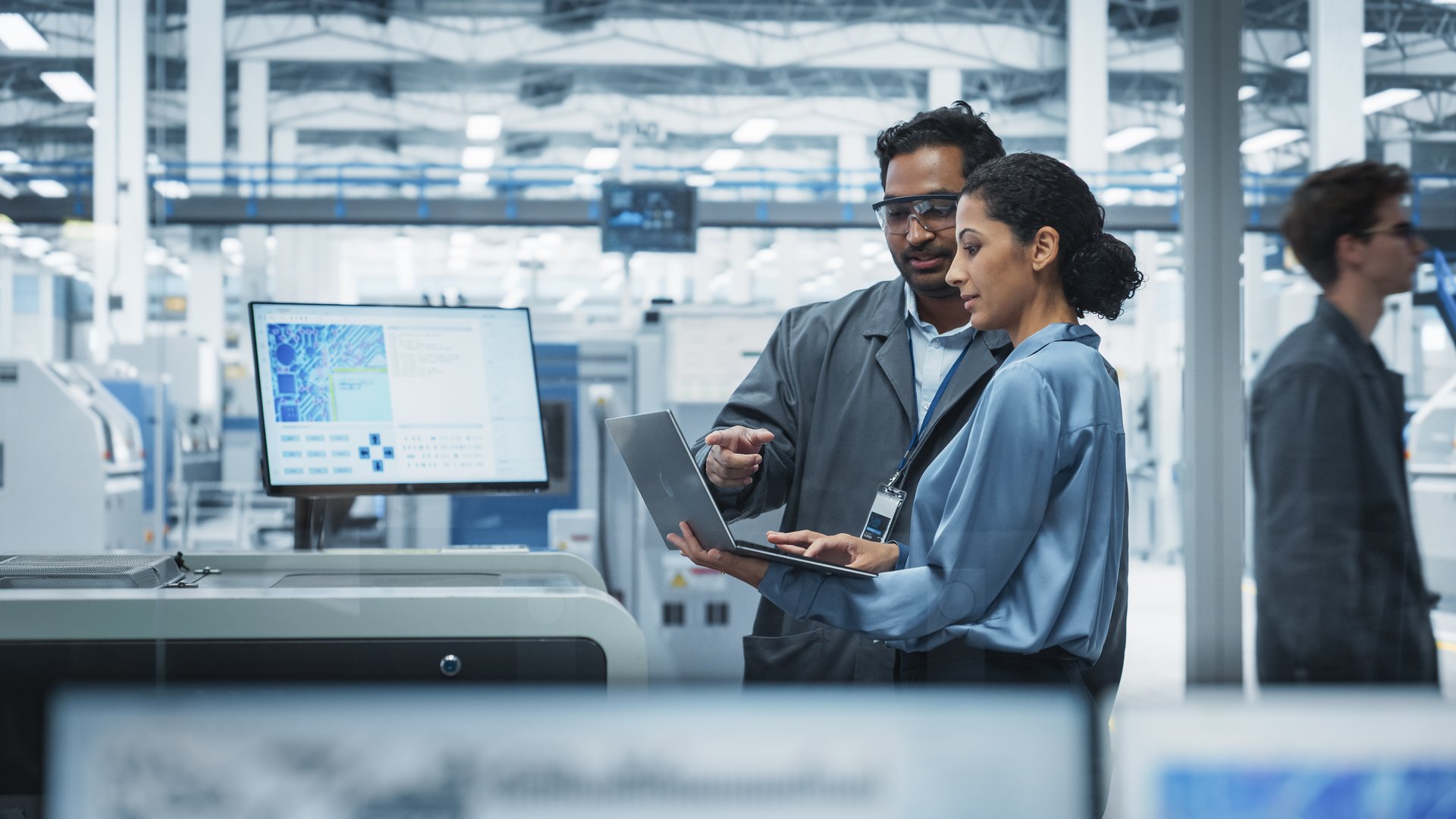 Indian Male Industrial Engineer And Hispanic Female Supervisor Using Laptop And Talking At An Electronics Factory.