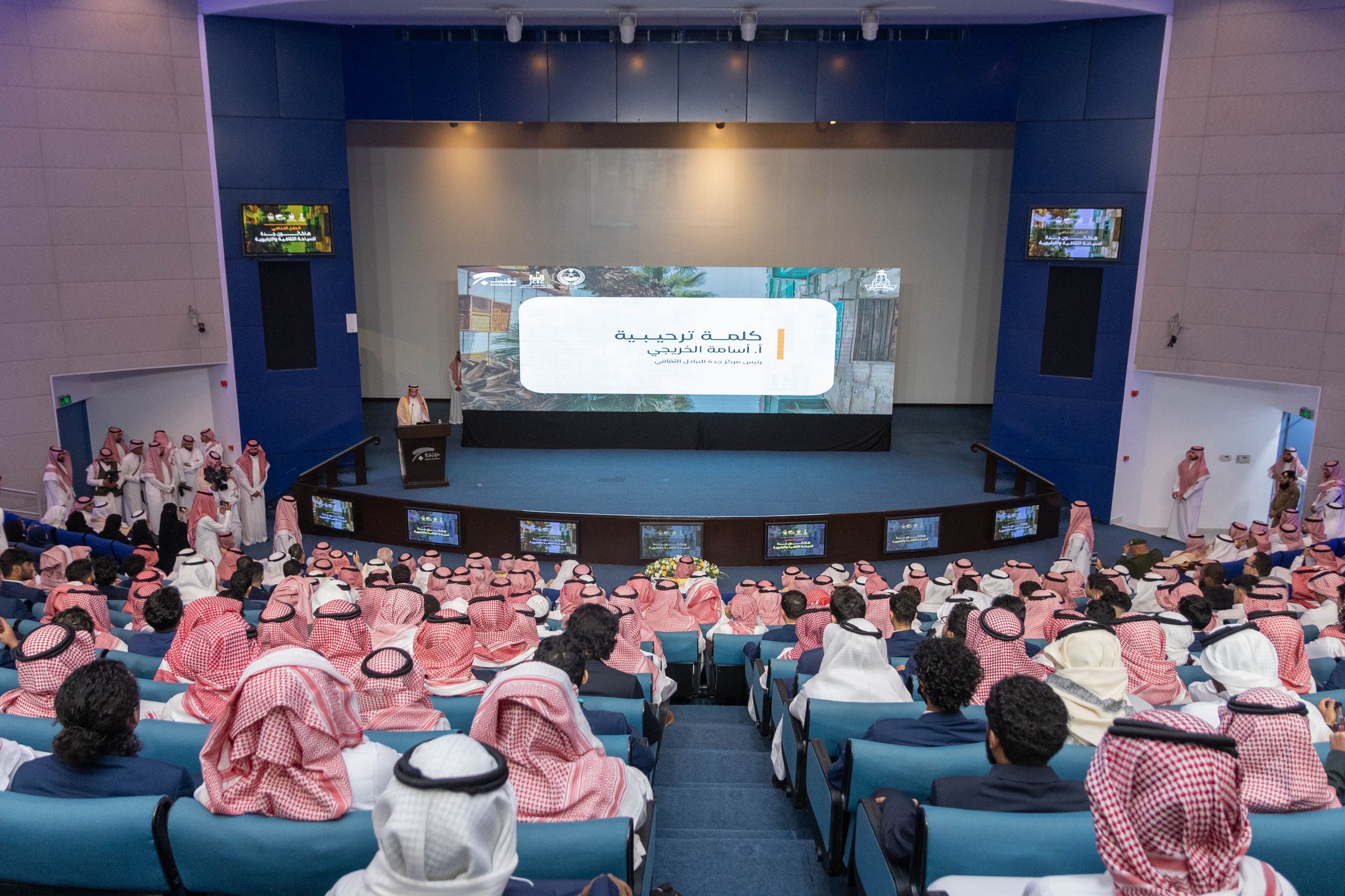 A conference room filled with attendees in traditional attire, focusing on a speaker at a podium and a large screen display.