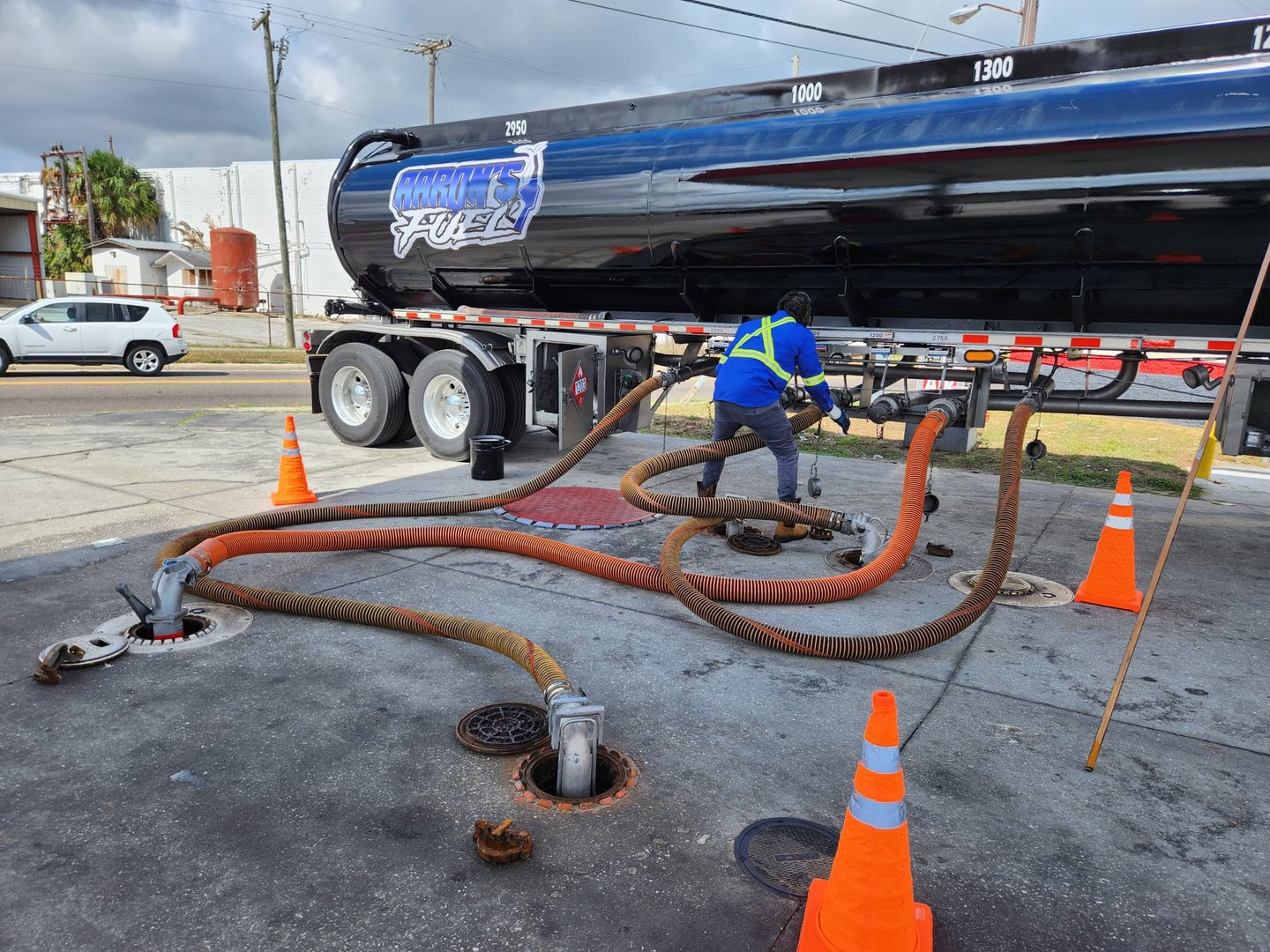 Worker in safety gear connects hoses to a large fuel truck at a gas station, surrounded by traffic cones.