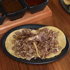 A plate with tacos filled with chopped grilled meat, placed on a wooden table with sauce containers in the background.