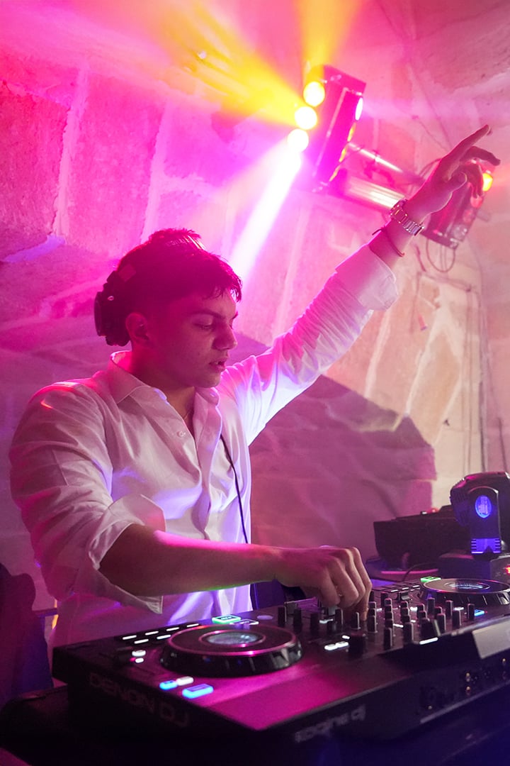 A DJ in a white shirt playing music on a console, with colorful lights and a stone wall background.