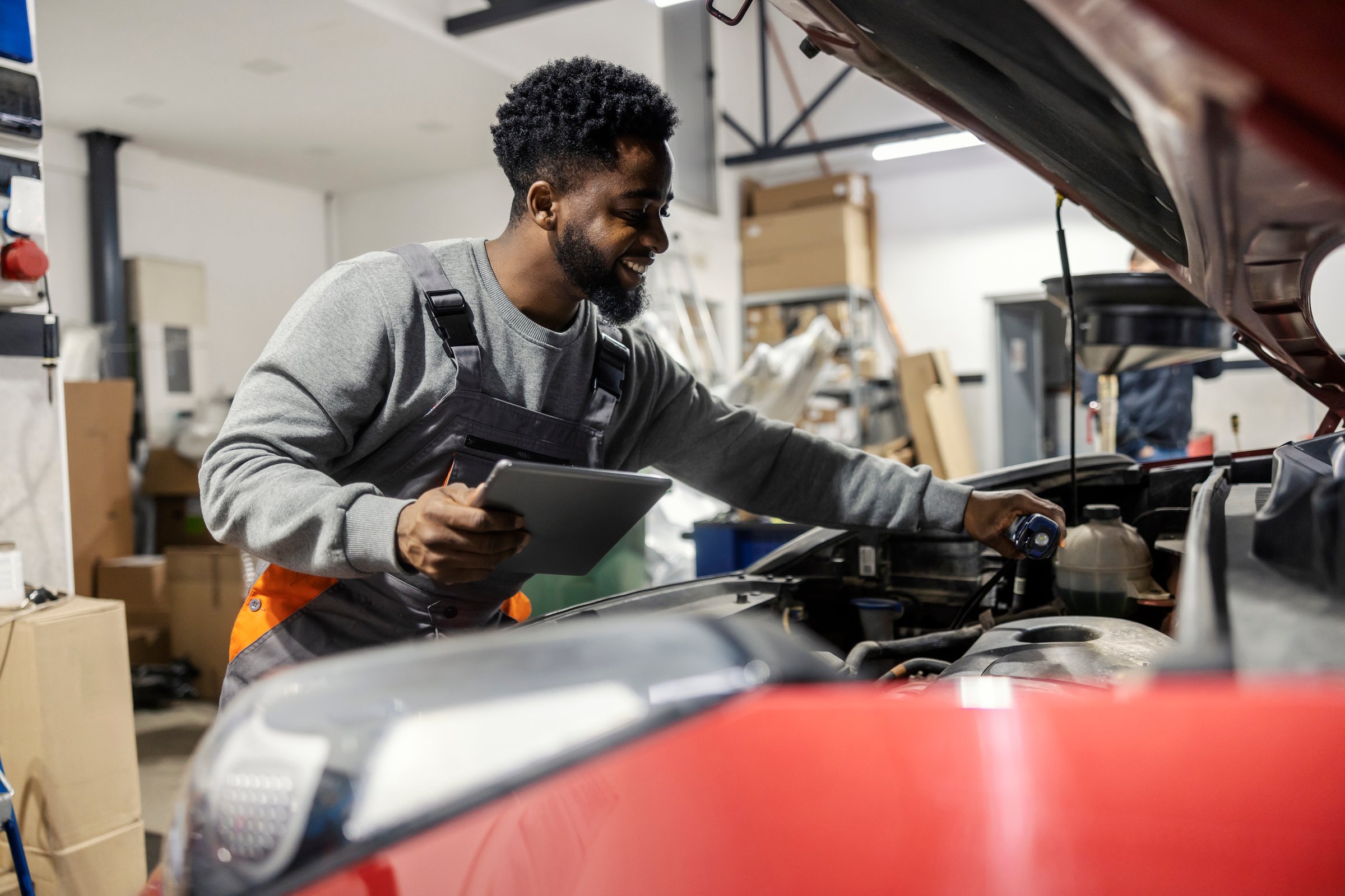 Smiling african american car service worker with tablet in hands pouring motor oil under car hood at automotive car service center.