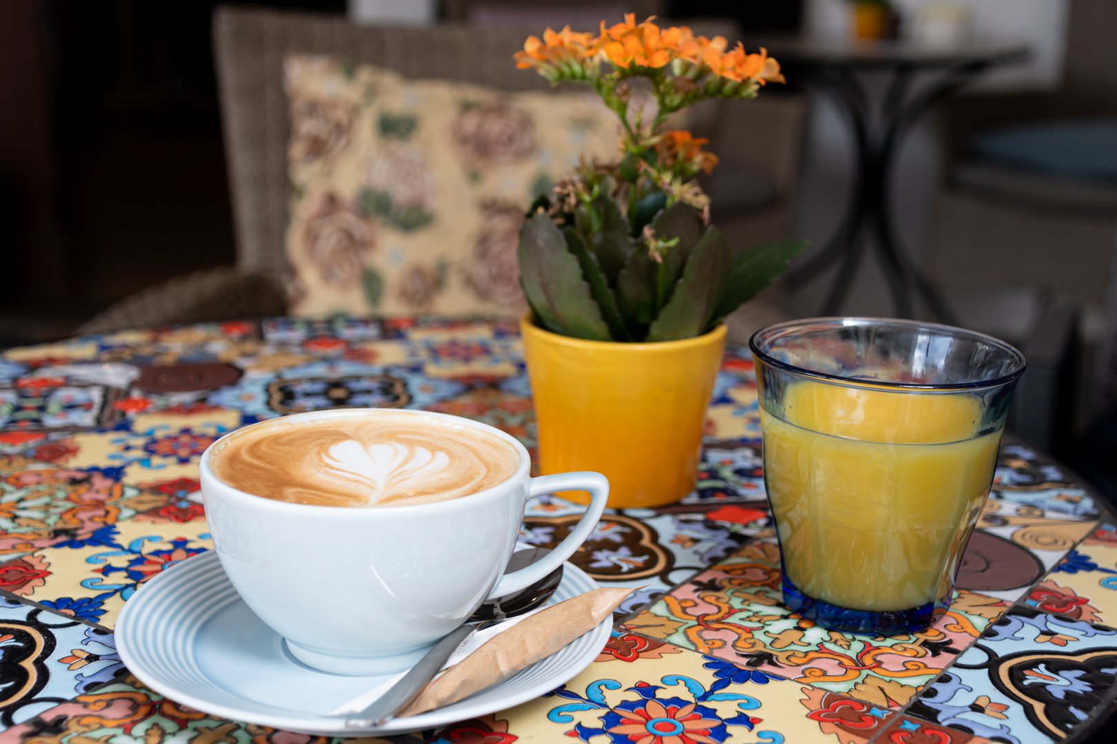 Beautiful and colorful table of different tiles, on which you can find a delicious cup of cappuccino coffee and fresh orange juice, as decoration, a vase. Beautiful decoration of a cozy place to have breakfast or coffee.