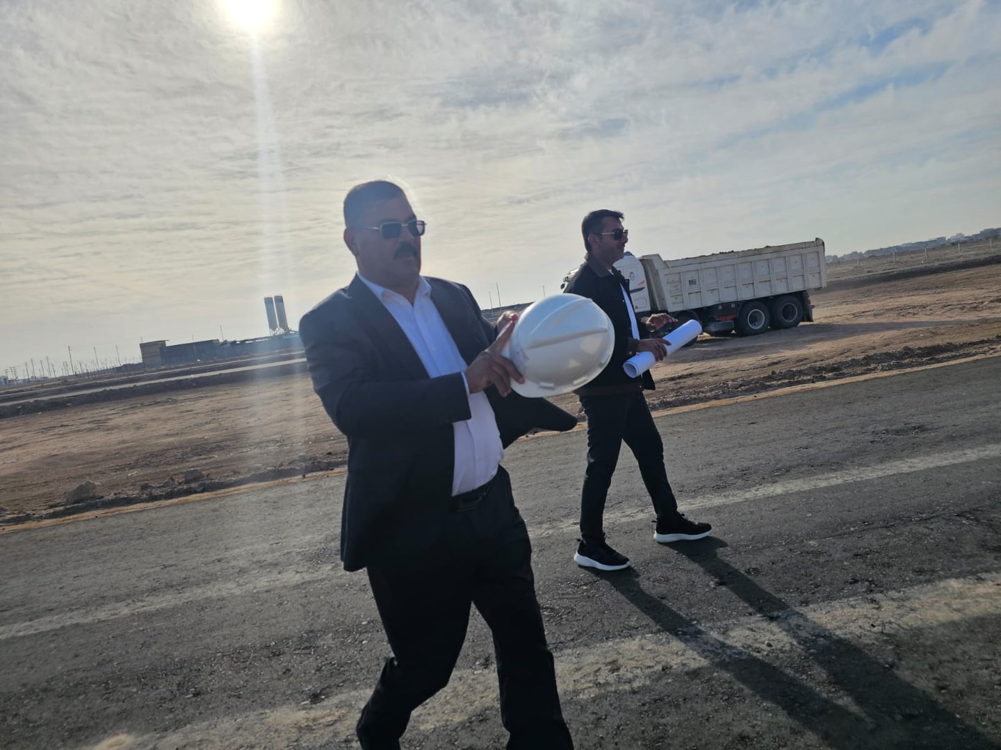 Two men in suits walking on a construction site, one holding a hard hat, with a truck and sunny sky in the background.