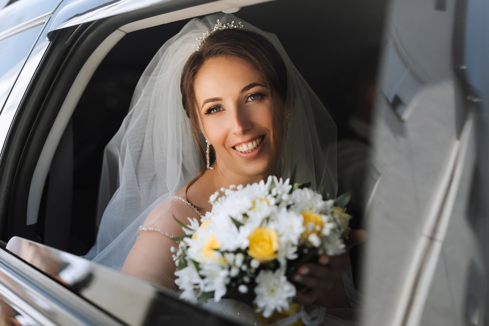 A bride is sitting in a car with a bouquet of flowers. She is smiling and looking out the window