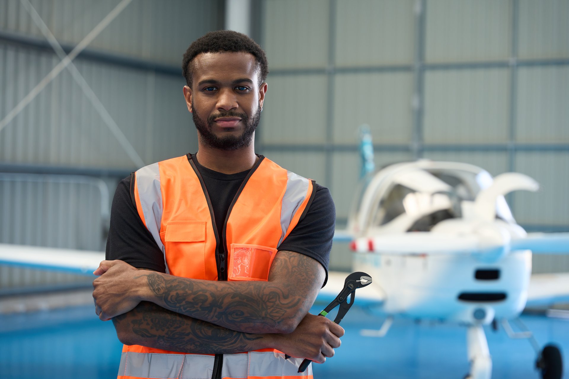 Confident aircraft mechanic holding pliers while posing in hangar with small passenger airplane in background