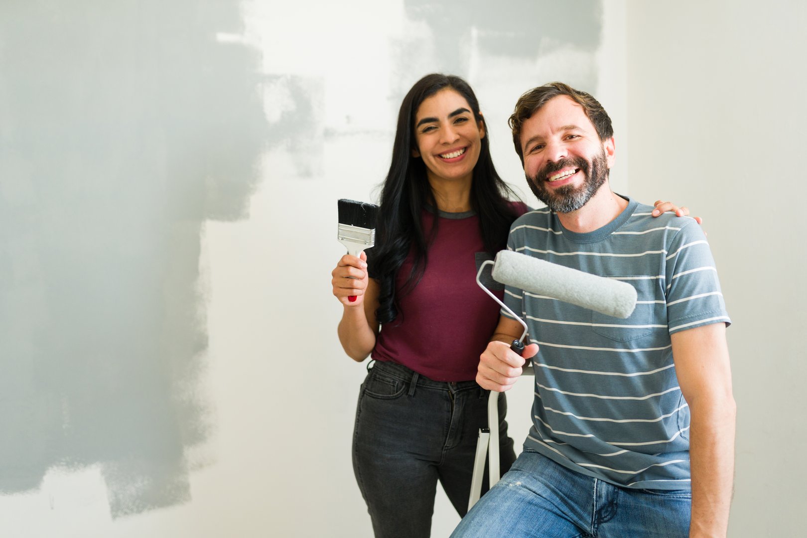 Happy couple holding paint rollers and brushes, taking a break while painting a wall gray in their new home